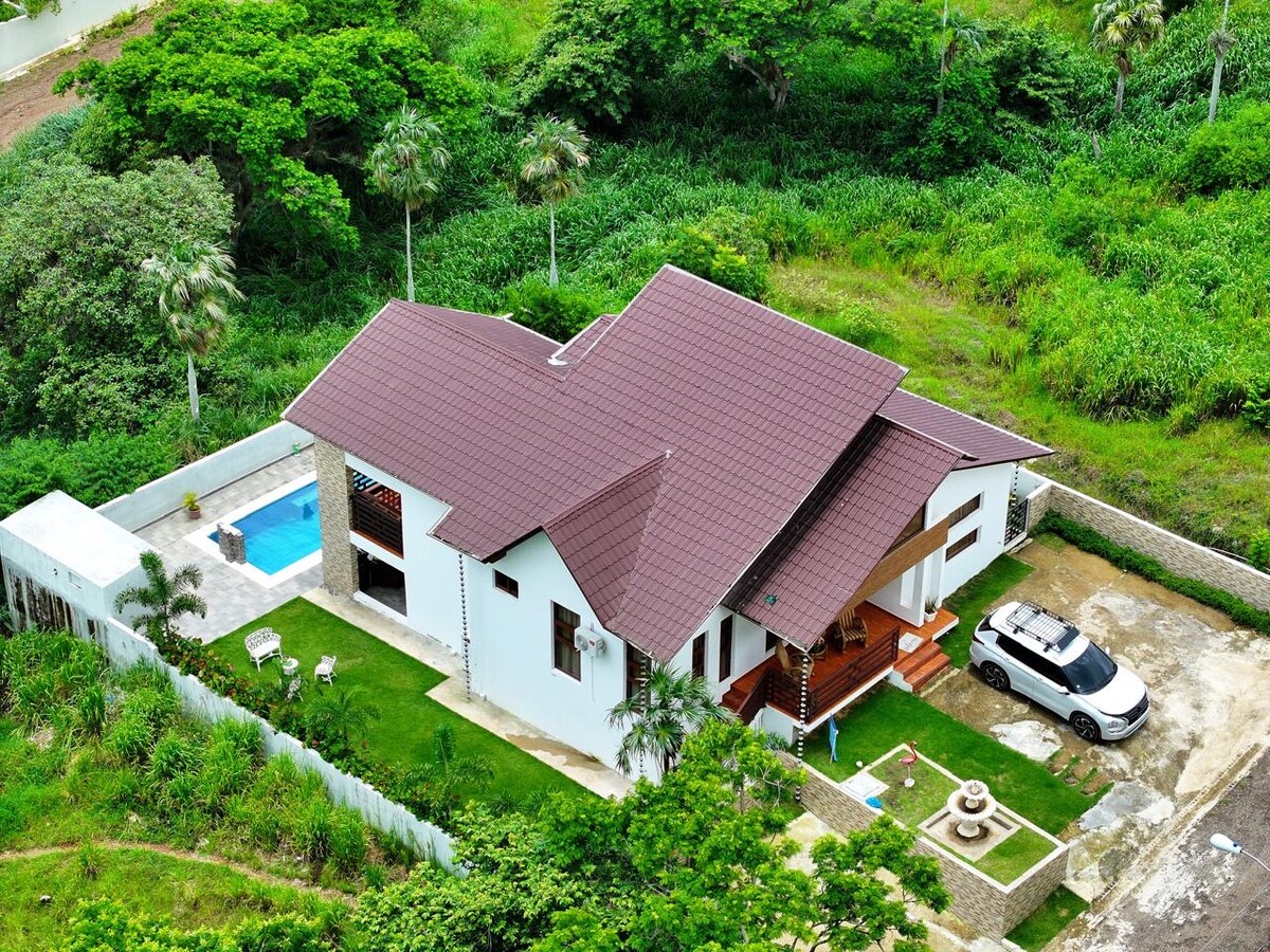 An aerial view showcases a spacious, modern home surrounded by lush greenery. A private pool can be seen beside the house, with a paved driveway leading up to the entrance. Outdoor seating is positioned on the lawn, emphasizing the tranquil surroundings.