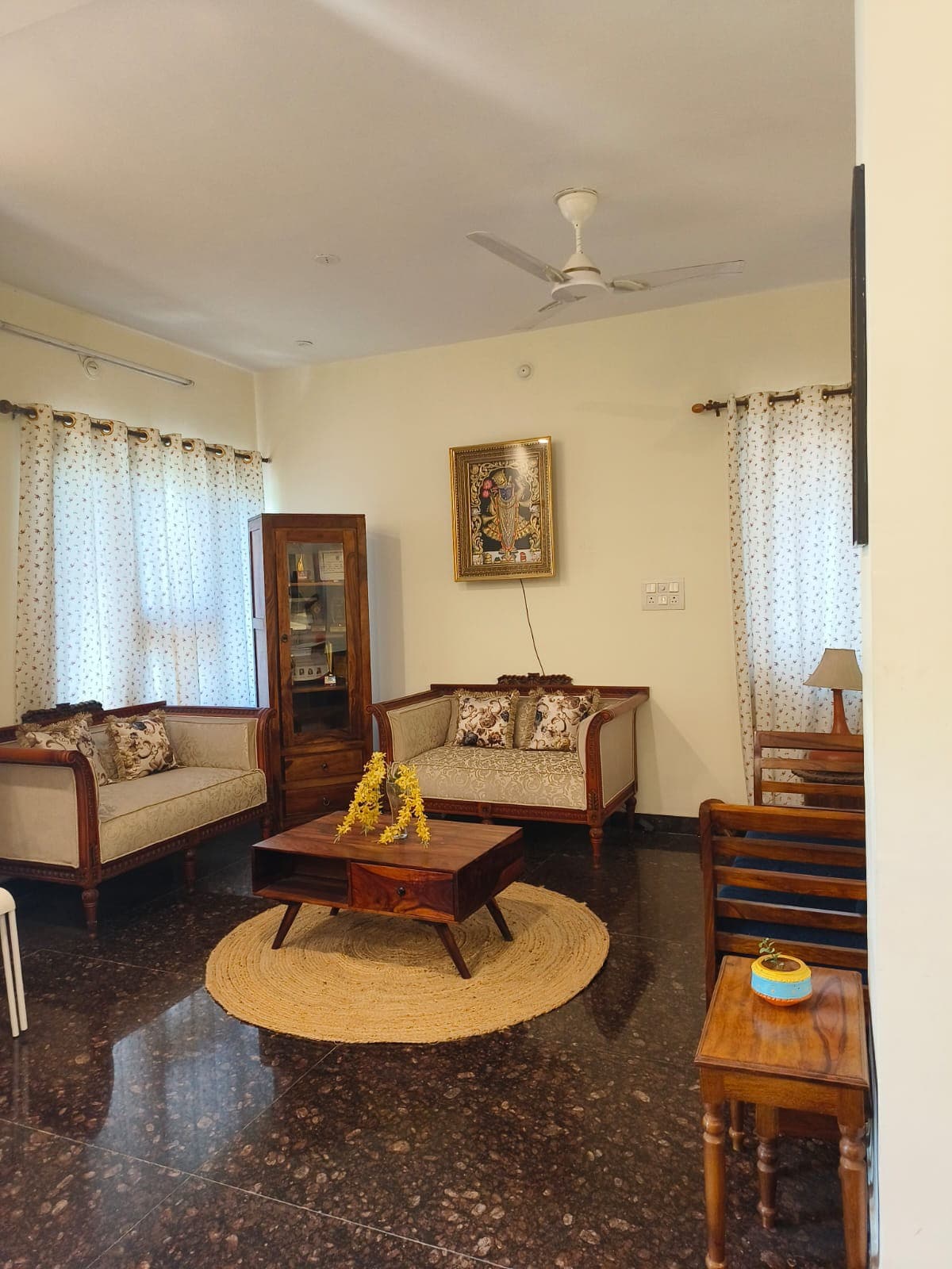 A cozy living area features two upholstered sofas adorned with decorative cushions, positioned around a central wooden coffee table. Natural light enters through large windows dressed in patterned curtains. A small lamp rests on a wooden side table, adding a touch of warmth to the space.