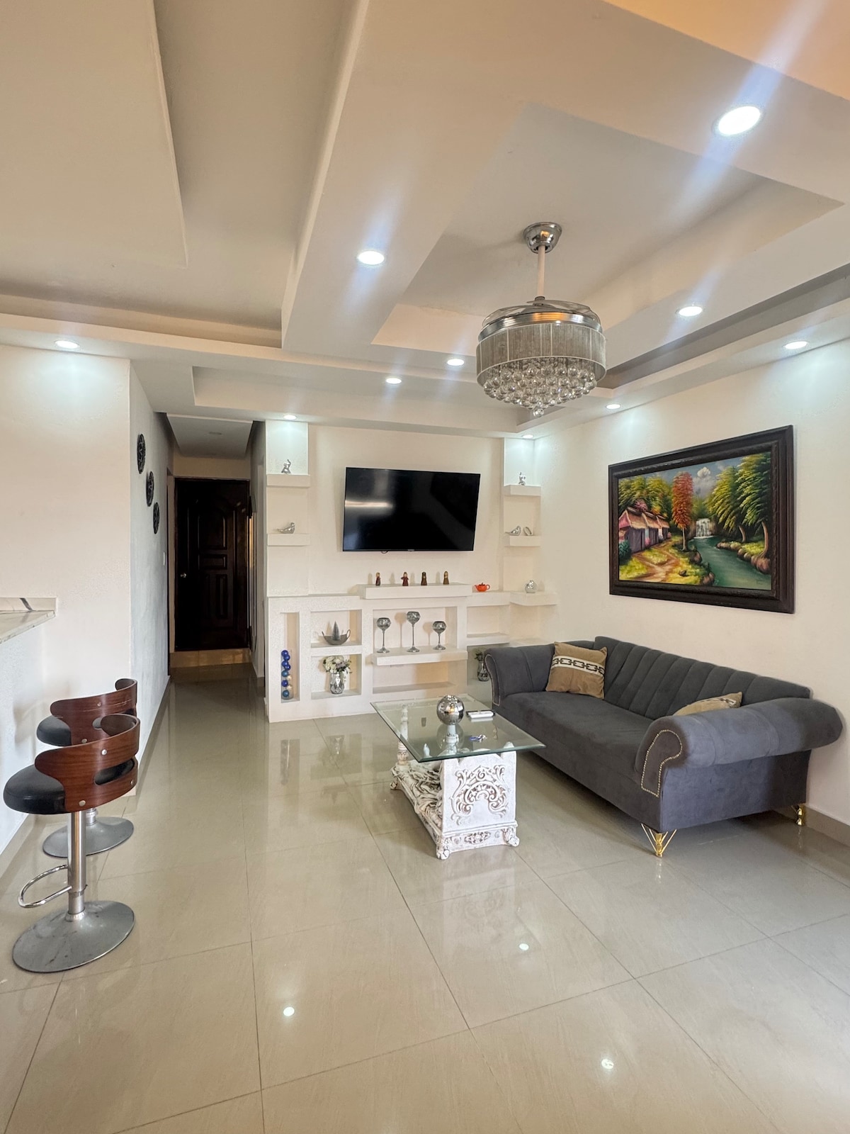 A modern living area is featured, showcasing a gray sofa paired with stylish décor. A light-colored coffee table stands in front of the sofa, while a large TV is mounted on the wall. Bright light illuminates the space from recessed ceiling fixtures.