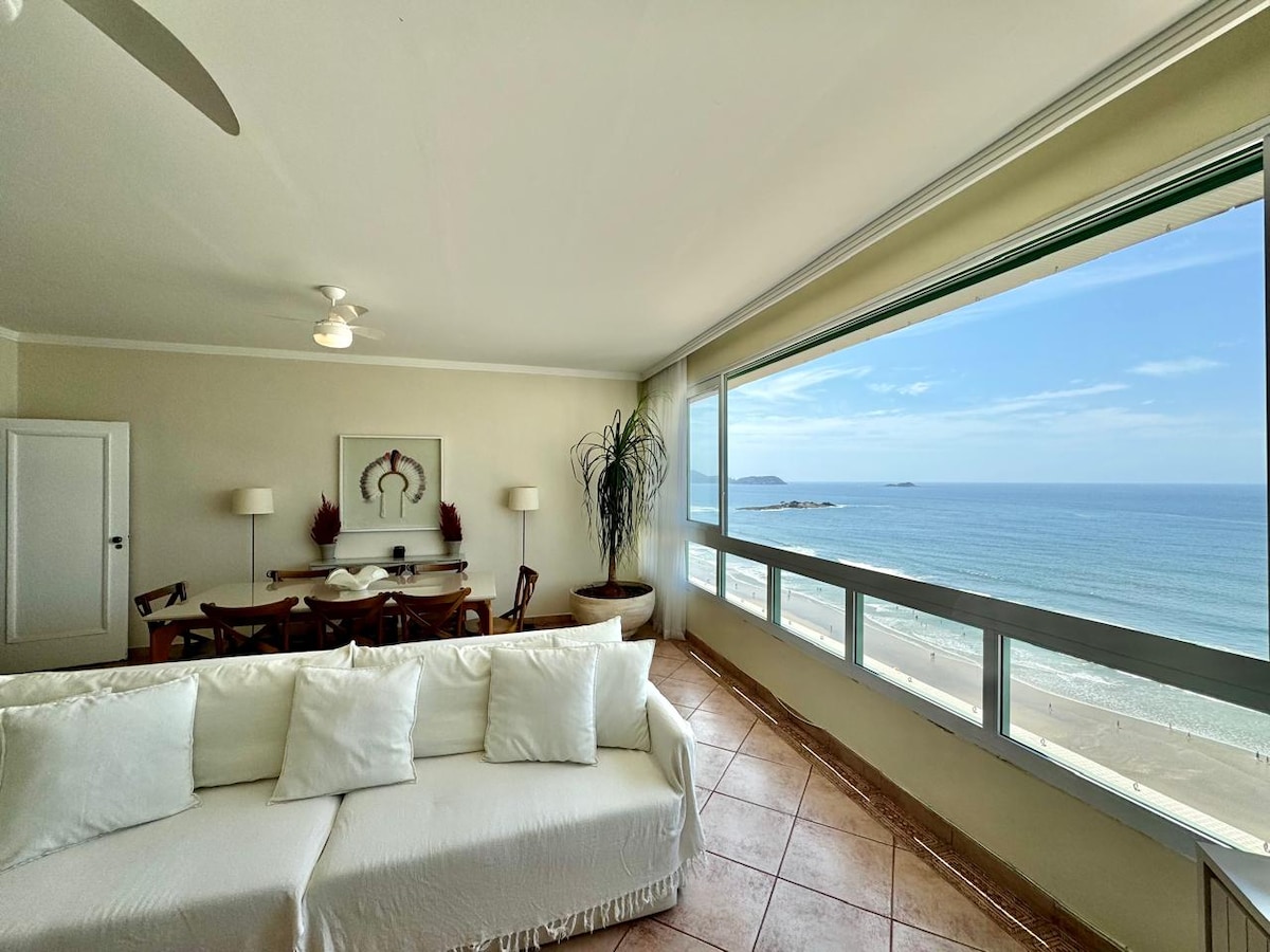 A bright living area is displayed, featuring a light-colored sofa facing large sliding glass doors that provide a clear view of the ocean. A dining table with wooden chairs is positioned in the background, contributing to a spacious and airy environment.