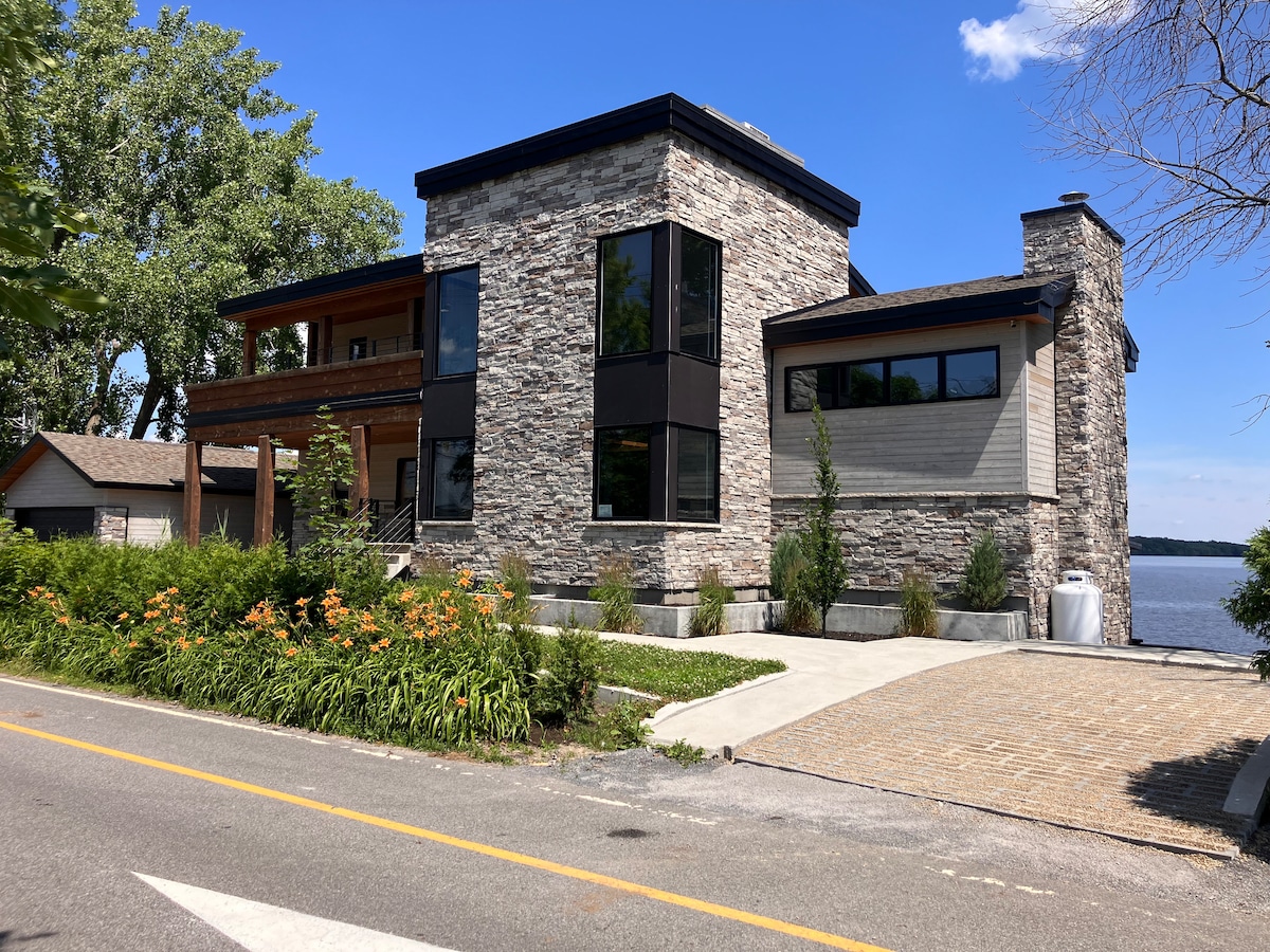 A modern stone-clad house is showcased against a clear blue sky. Lush greenery lines the pathway leading to the entrance, while large windows reflect the surrounding landscape. A vehicle parking area is visible, along with a waterfront view in the background.
