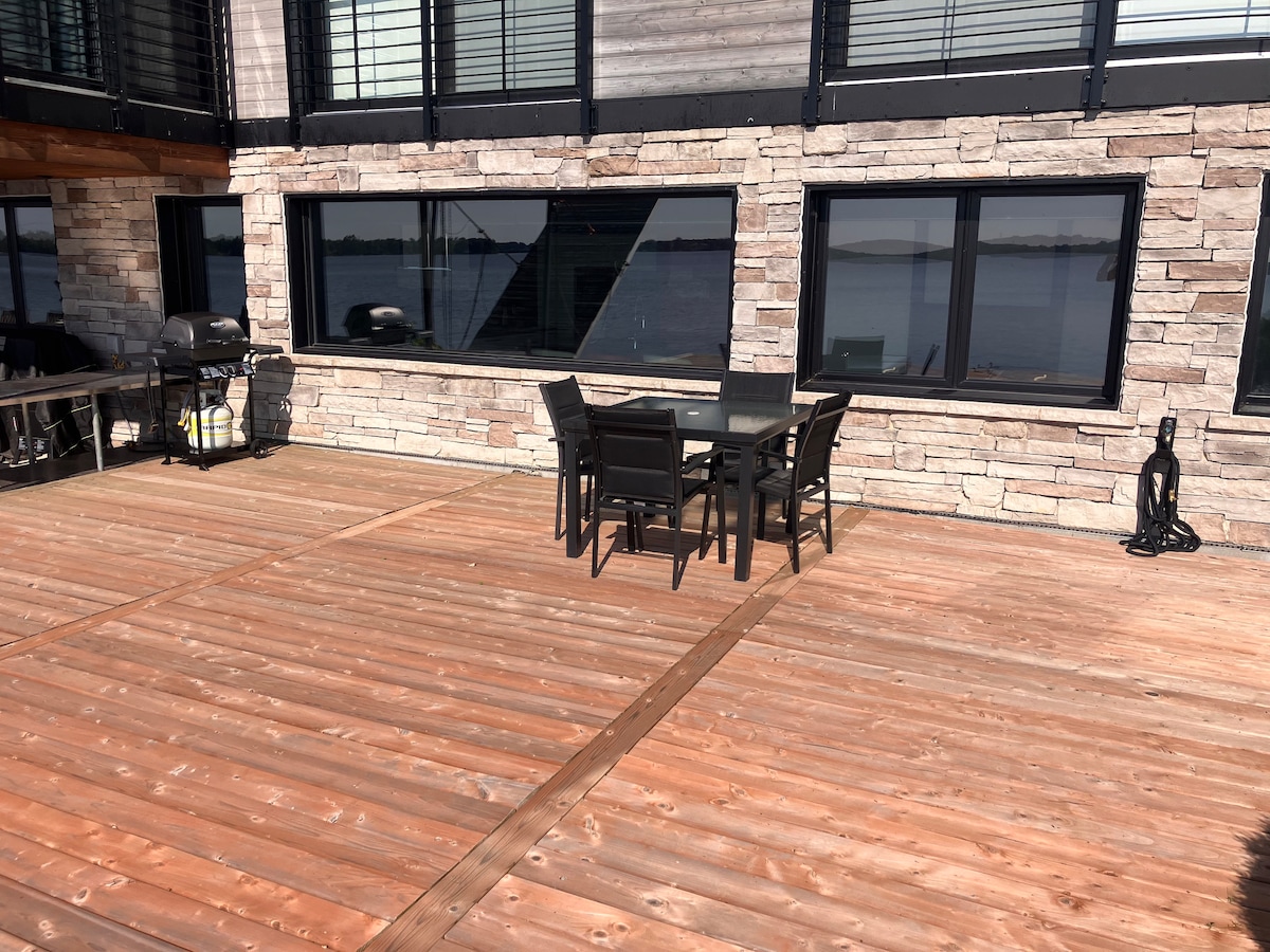 An outdoor wooden deck features a dining table and six chairs, positioned against a stone wall. Large windows with black frames offer views of the nearby water, reflecting natural lighting across the space. Grilling equipment is visible nearby.