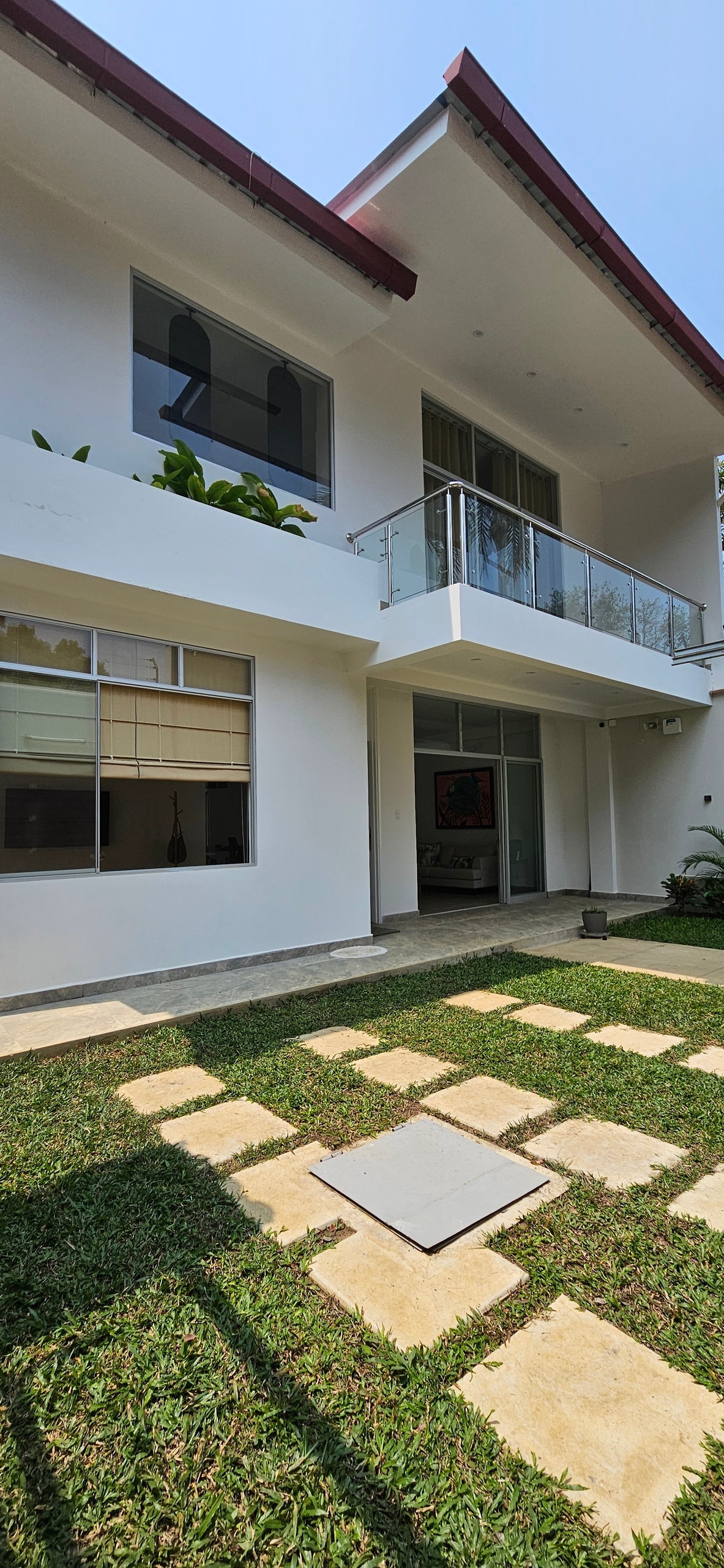 A two-story house is shown with large windows and a balcony. A neatly arranged stone pathway leads through a green lawn, complementing the white exterior walls. Potted plants are visible near the entrance, enhancing the inviting ambiance of the outdoor space.
