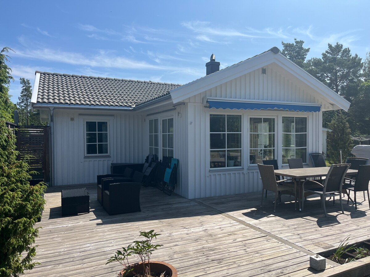 A charming villa with a light-colored exterior and multiple large windows is showcased. A spacious wooden deck is present, furnished with a dining table and comfortable seating areas. Surrounding greenery adds a natural touch, enhancing the inviting outdoor space.