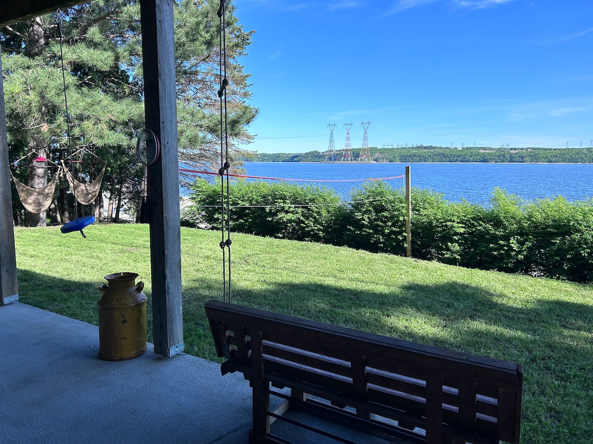 A covered patio offers a relaxing view of the expansive water, framed by lush greenery. A rustic wooden bench is positioned invitingly, and a vintage milk can adds charm to the space. Hammocks are strung nearby, providing spots for relaxation.