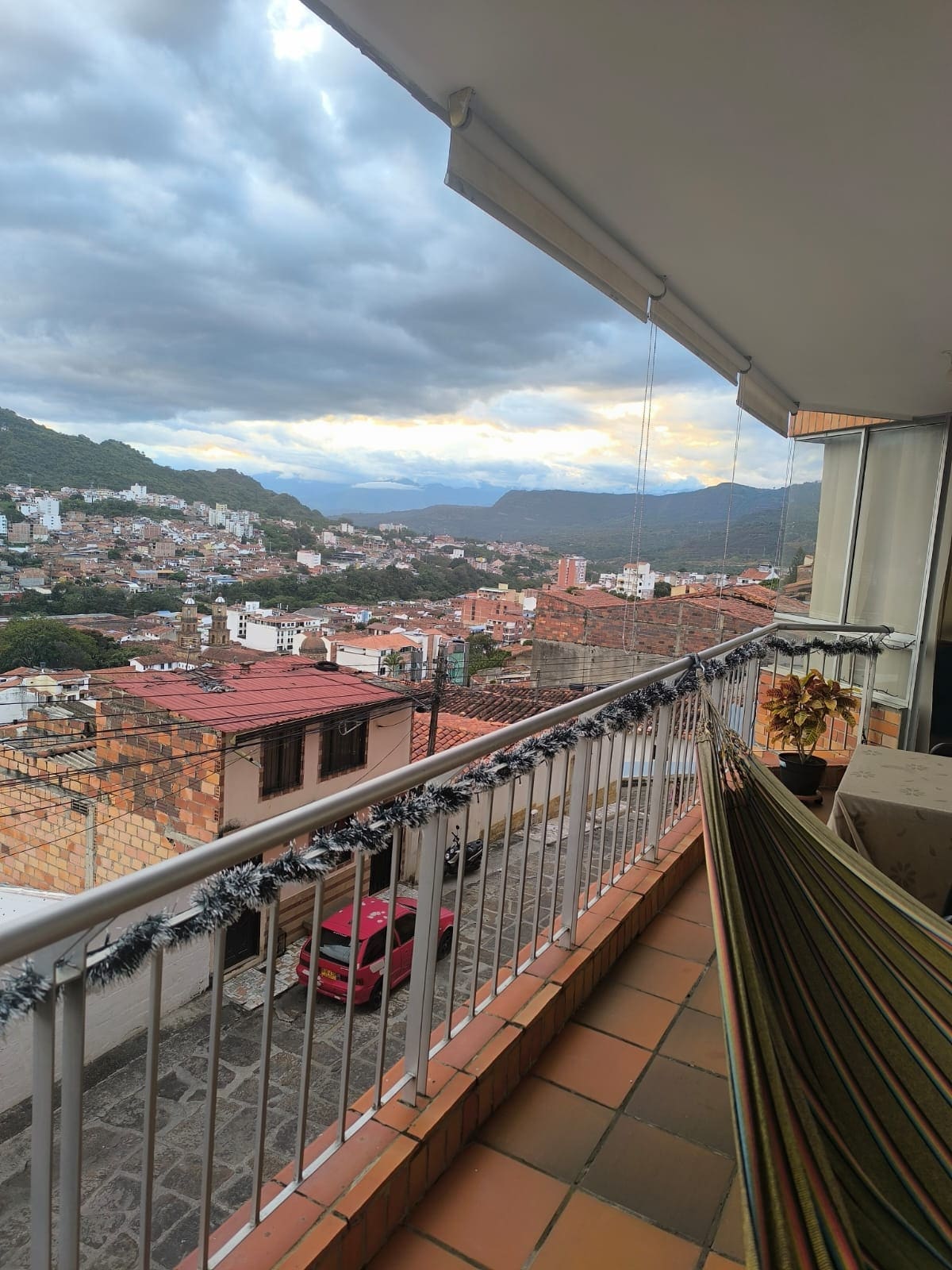 A spacious balcony is featured, showcasing a view of the surrounding mountains and town. A comfortable hammock hangs in the foreground, while cozy seating arrangements are visible. The exterior is adorned with decorative elements, enhancing the inviting outdoor space.