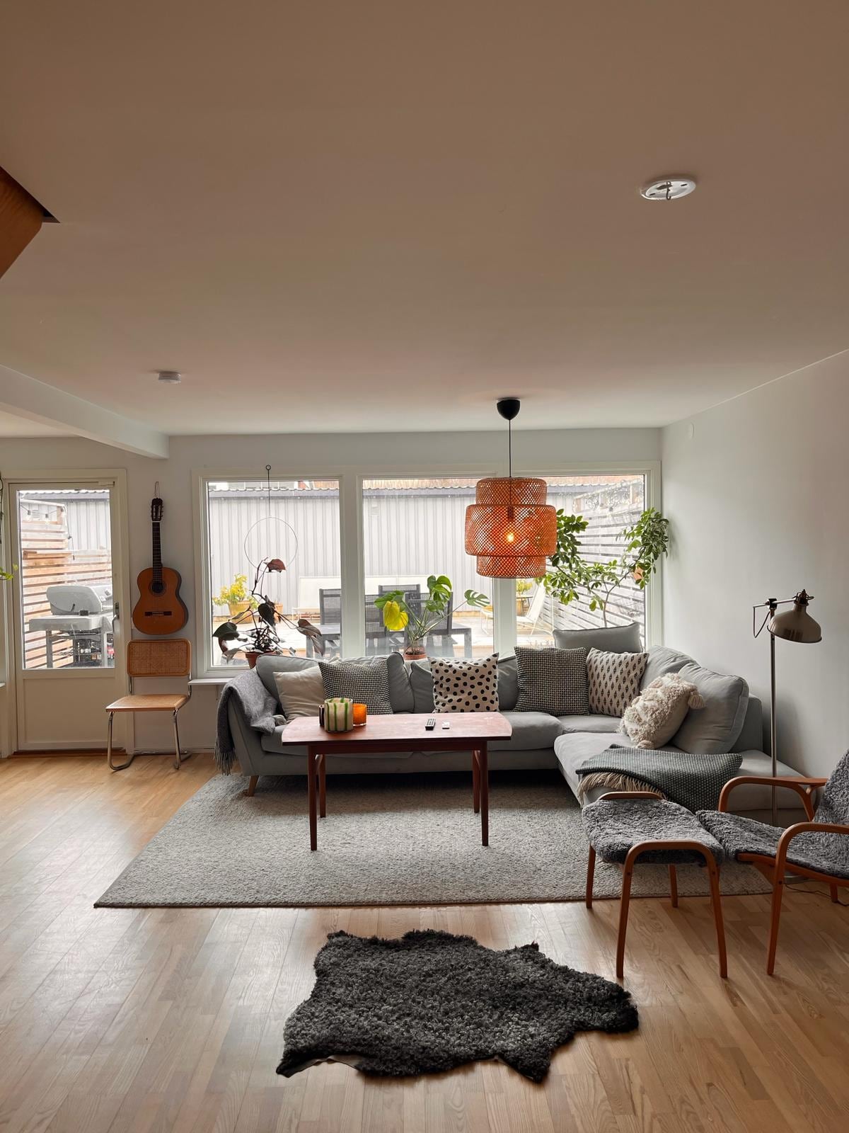 A cozy living room features a comfortable gray sofa adorned with patterned cushions. A coffee table sits on a soft rug, and a guitar is present in the corner. Light enters through large windows, complemented by indoor plants and warm lighting in the form of a pendant lamp.