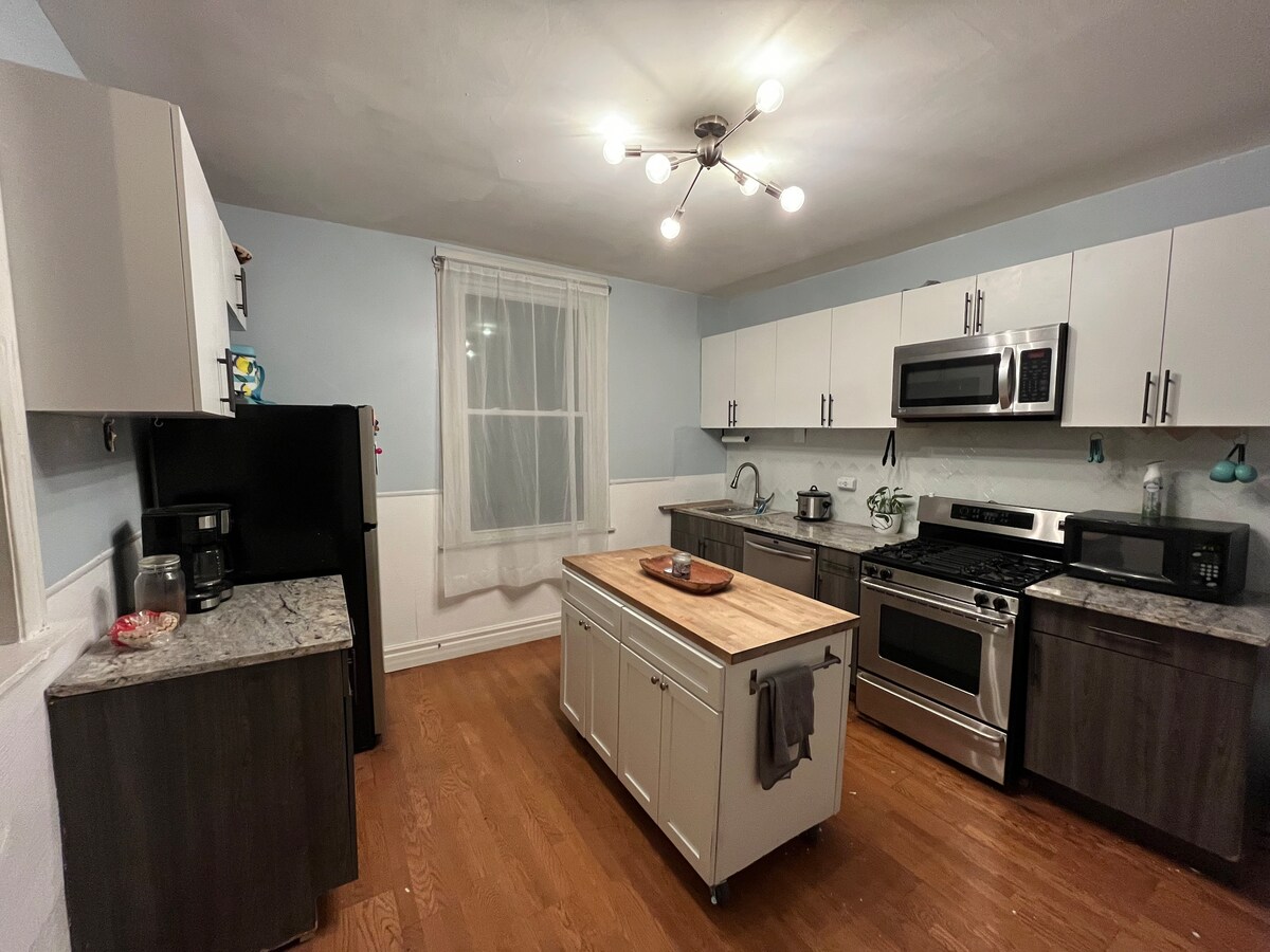 The kitchen features modern cabinetry with a light and dark color scheme. A central island with wooden countertop provides additional workspace. Stainless steel appliances, including a gas stove and microwave, are visible. Natural light enters through a large window, illuminating the room.