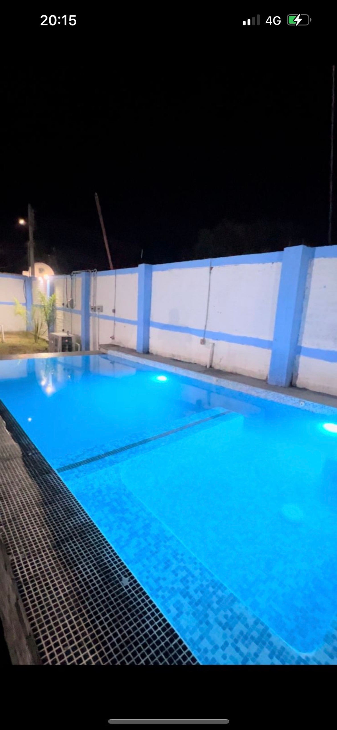 A serene night view of a swimming pool featuring bright blue waters, illuminated by underwater lighting. The enclosed area is surrounded by white walls, providing privacy and a sense of seclusion. Reflections from the pool add to the tranquil atmosphere.