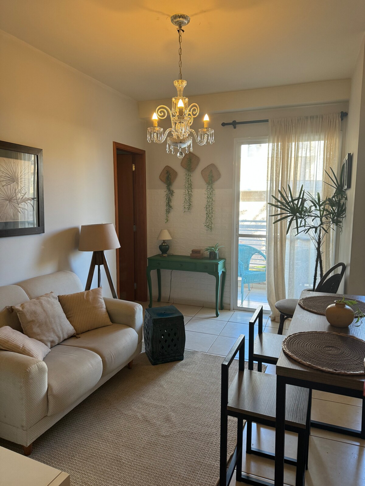 A cozy living area features a light-colored couch with decorative pillows, accompanied by a small table and chair set. Natural light enters through a glass door that leads to a balcony, accented by potted plants and wall decor. A chandelier hangs from the ceiling.