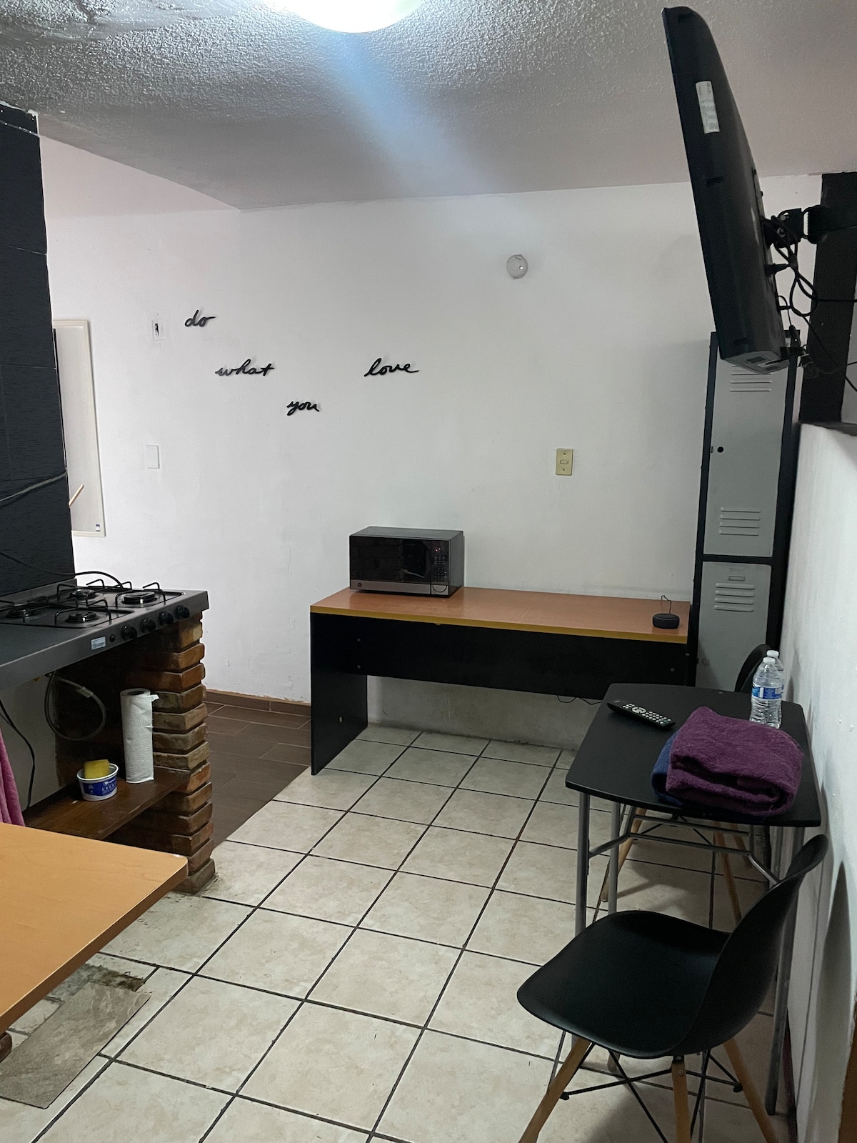 A compact kitchenette area is featured, including a small microwave and a workspace with a desk and chair. The walls are adorned with minimalistic text. A brick accent adds warmth to the tiled flooring, and a television is mounted near the workspace.