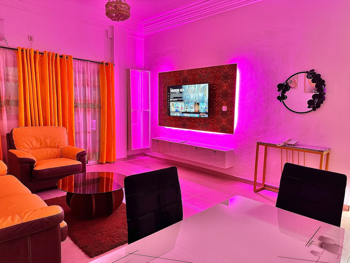 A vibrant living room features soft seating in warm shades beside a round glass coffee table. A smart TV is mounted on the wall, displaying a streaming service. Curtains of bright orange enhance the atmosphere, and decorative lighting creates an inviting ambiance.