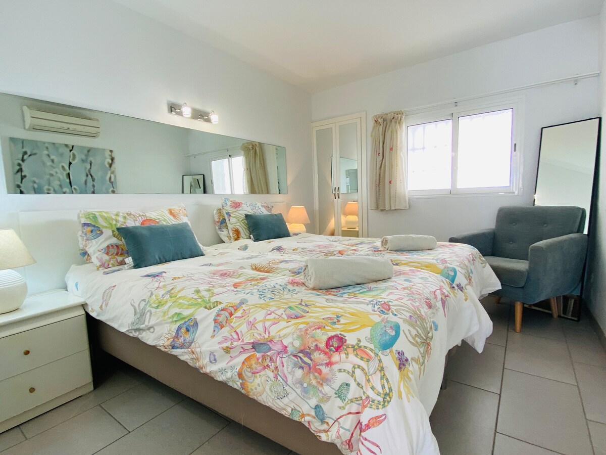 A well-appointed bedroom is presented with a spacious bed featuring a colorful patterned bedspread. Natural light filters through the window, brightening the room. A mirrored wall adds depth, while a corner chair and bedside lamps enhance the inviting appearance of the space.