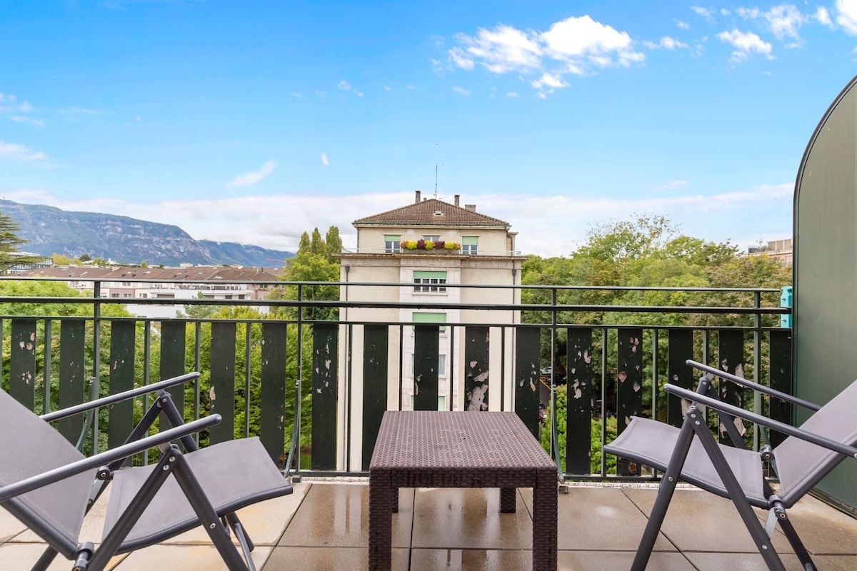 A south-facing terrace features two modern folding chairs and a small table, set against a backdrop of green trees and distant mountains. The open sky allows ample natural light, creating an inviting space for relaxation.