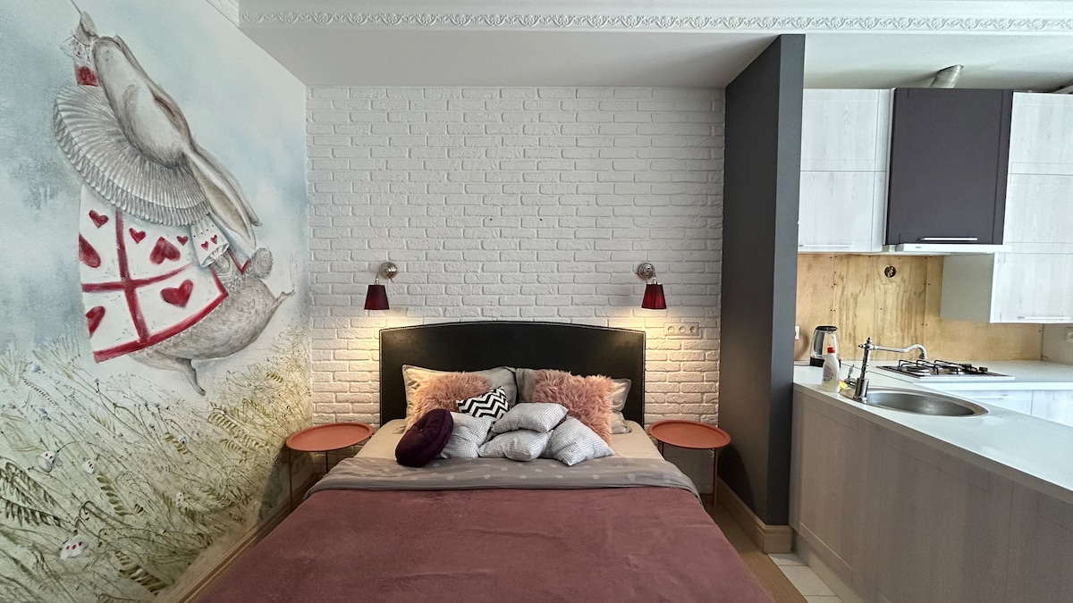 A compact studio features a unique wall mural depicting a whimsical character alongside a white-brick feature wall. The spacious bed is adorned with soft cushions and placed between two small bedside tables, while a modern kitchen area is visible in the background.
