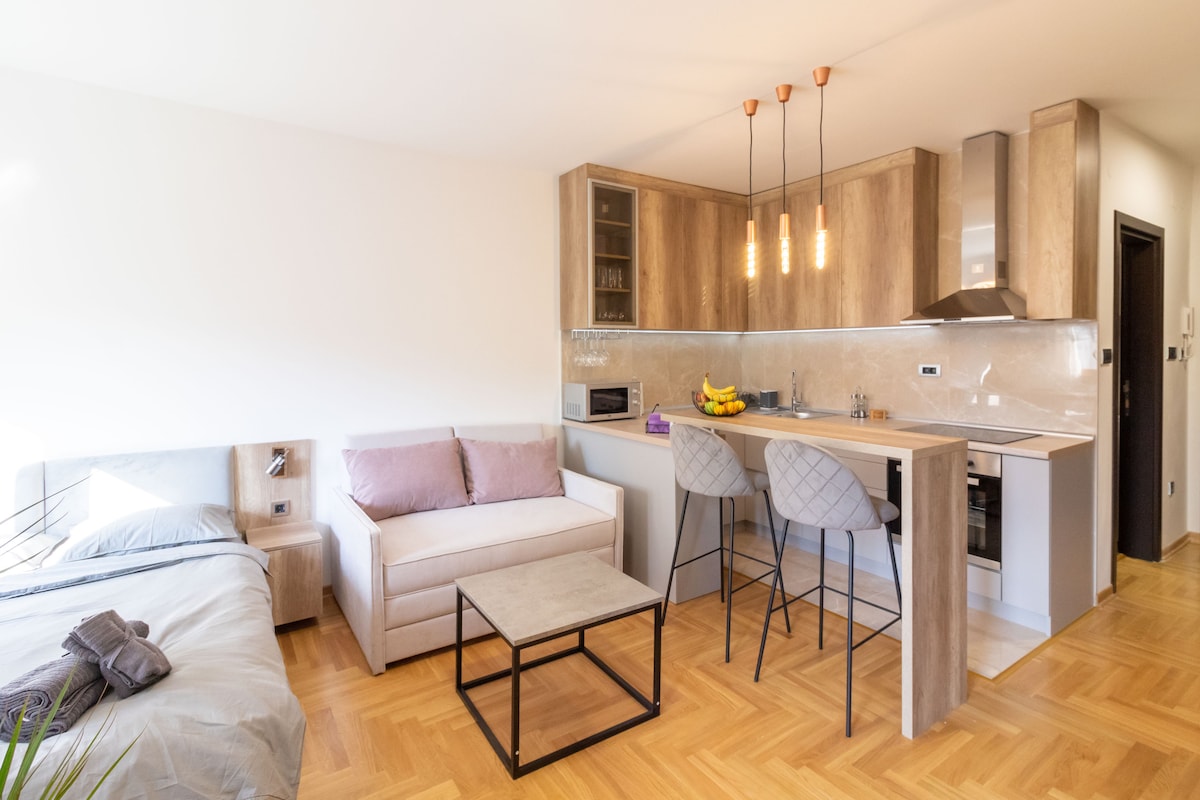 A modern living space showcases an open layout, featuring a cozy sleeping area with a neatly made bed beside a stylish sofa. A rectangular coffee table sits centrally, while bar stools are positioned at a sleek kitchen counter with overhead pendant lighting.