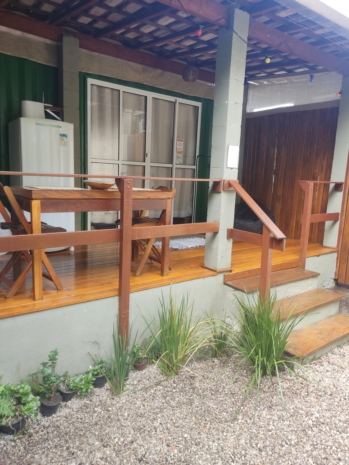 A porch area is featured, with a wooden deck leading to steps. A dining table with chairs is situated on one side, while a refrigerator is visible behind a glass door. Lush green plants line the edge of the gravel pathway.