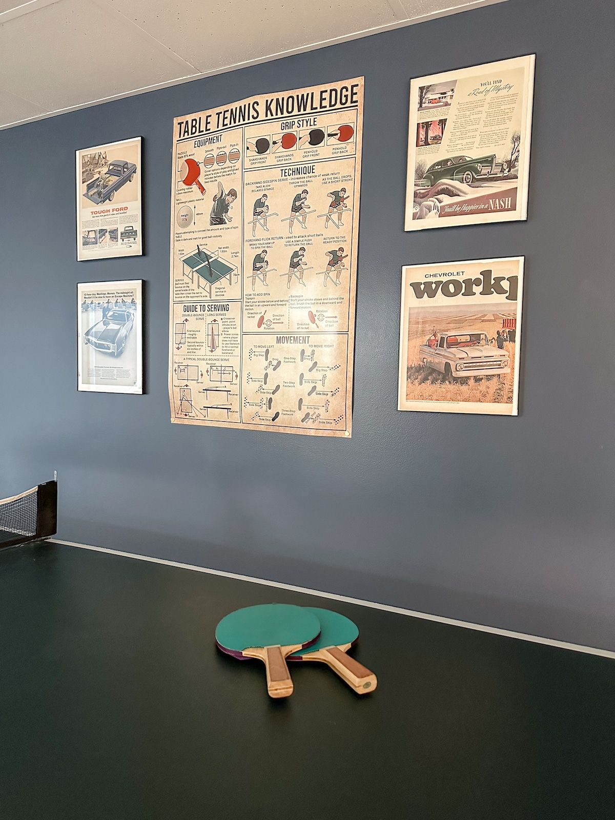 A dedicated game area features a ping pong table with a green playing surface. Two paddles rest beside the table. Vintage posters adorn the deep blue walls, displaying illustrations and advertisements related to cars and table tennis.