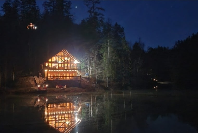The cabin is illuminated at night, reflecting beautifully on the still waters of Spirit Lake. Tall trees surround the property, and soft lights accentuate the cabin's architecture, creating a serene ambiance. Outdoor seating can be seen near the water's edge.