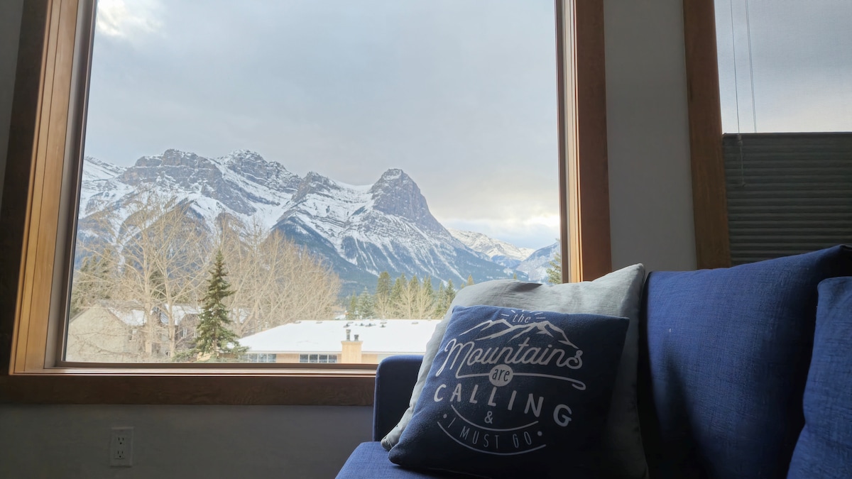 A large window offers a view of the snow-capped mountains in the distance. A blue couch with a mountain-themed pillow sits nearby, inviting relaxation while taking in the scenic landscape.