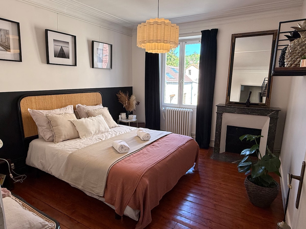A serene bedroom is presented with a spacious bed featuring light bedding and decorative pillows. Natural light enters through a window, enhancing the overall ambiance. Wall art is displayed above the bed, and a large mirror reflects elements of the room, including a potted plant adjacent to the fireplace.