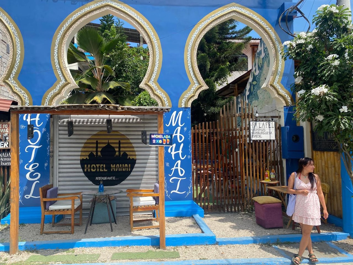 The hotel entrance is characterized by vibrant blue walls and decorative arches. A small seating area with two chairs is positioned next to the entrance. Lush greenery and flowers are integrated into the surrounding landscape, enhancing the inviting atmosphere of the hotel.