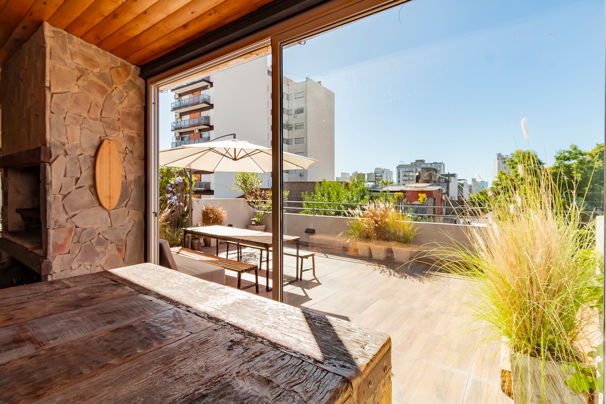 A spacious terrace is seen through large glass doors, featuring a wooden dining table and an umbrella for shade. Lush greenery adorns the area, adding a natural touch. The terrace is bathed in sunlight, offering a vibrant outdoor space for relaxation.