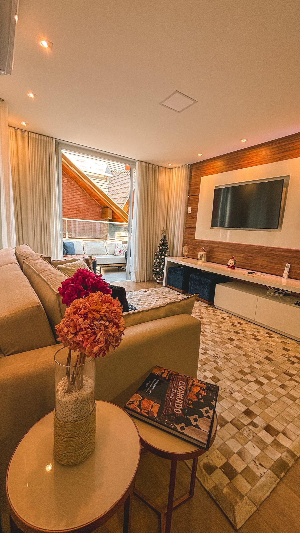 The living room features a beige sofa positioned across from a mounted flat-screen TV. A decorative rug covers the floor, while natural light is brought in through large windows adorned with sheer curtains. A small table holds a vase of flowers and magazines.