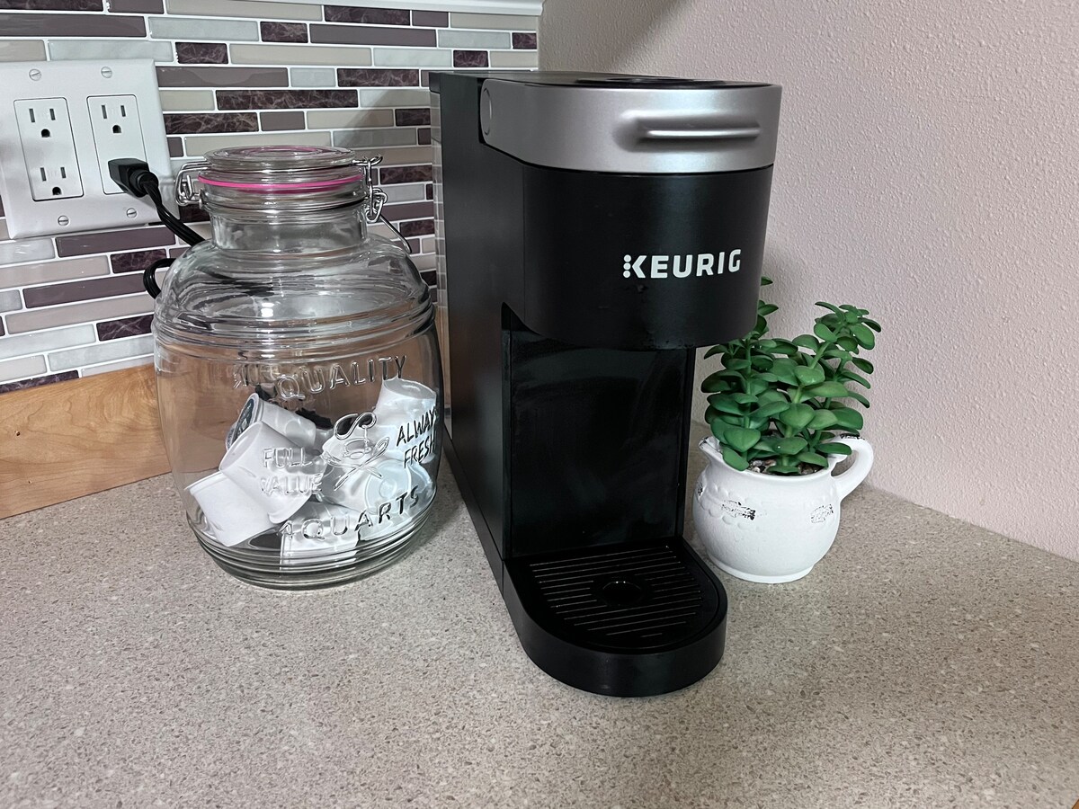 A countertop hosts a black Keurig coffee maker beside a glass jar filled with sugar packets. A small potted plant adds a touch of greenery, complementing the mosaic backsplash that enhances the kitchen's modern design.