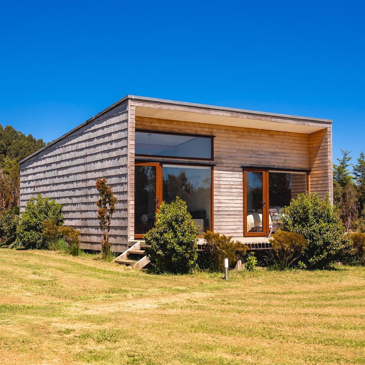 This modern cabin features a wooden exterior and large windows that allow natural light to fill the interior. Surrounding greenery and a well-maintained lawn create a serene outdoor environment, enhancing the connection with nature.