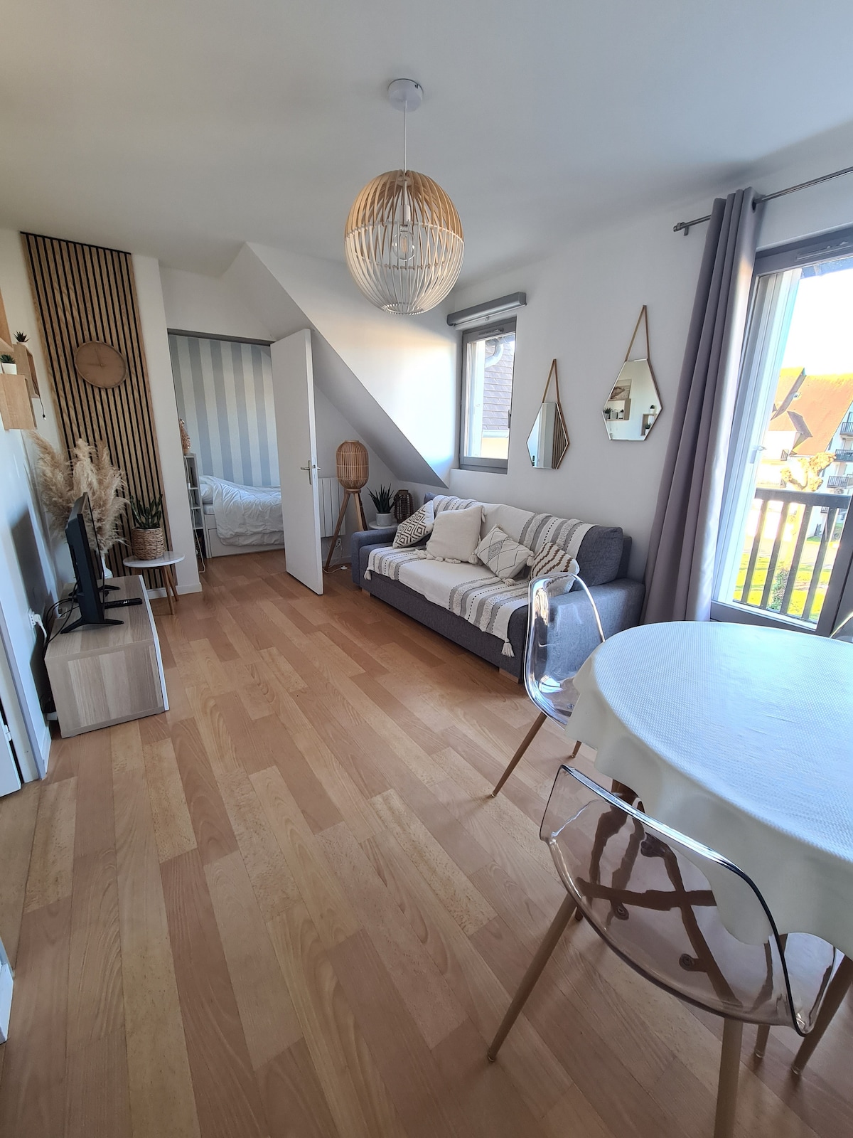 A bright, airy living space is visible, featuring a striped sofa and a TV. An adjacent dining area is indicated with a round table and transparent chairs. Natural light is permitted through large windows, which overlook a private balcony. A bedroom entrance is partially seen in the background.