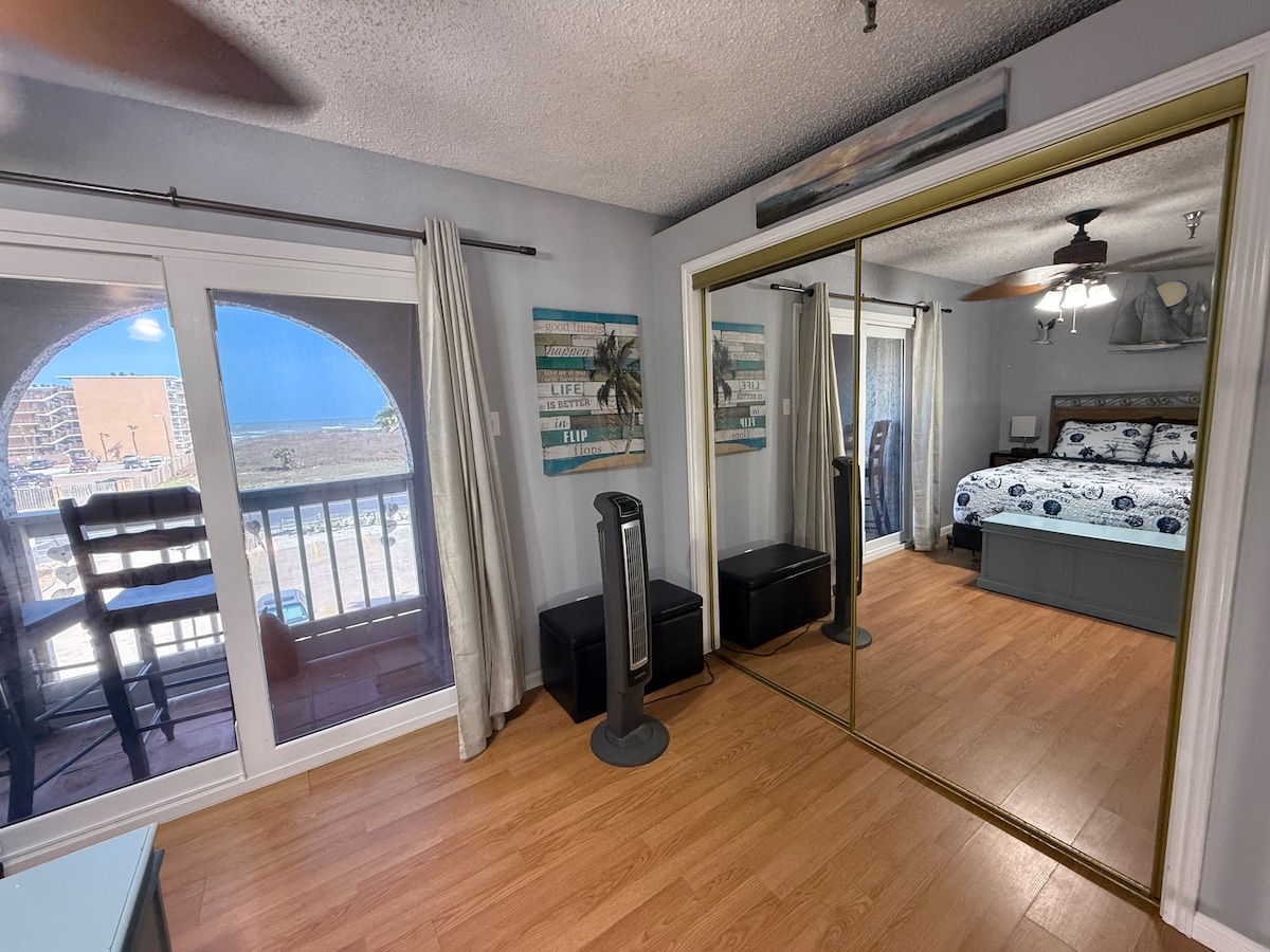A spacious loft bedroom features a queen size bed with beach-themed linens. Natural light enters through sliding glass doors, leading to a balcony with a view. A mirrored closet reflects the room's calming decor and wood-style flooring. Practical items like a fan and storage boxes are also present.