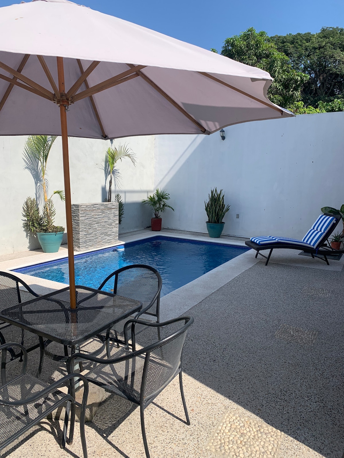 A private pool is positioned within a secluded patio, surrounded by potted greenery. A shaded table with four chairs offers a space for relaxation, while a striped lounge chair rests nearby, inviting enjoyment of the warm weather.
