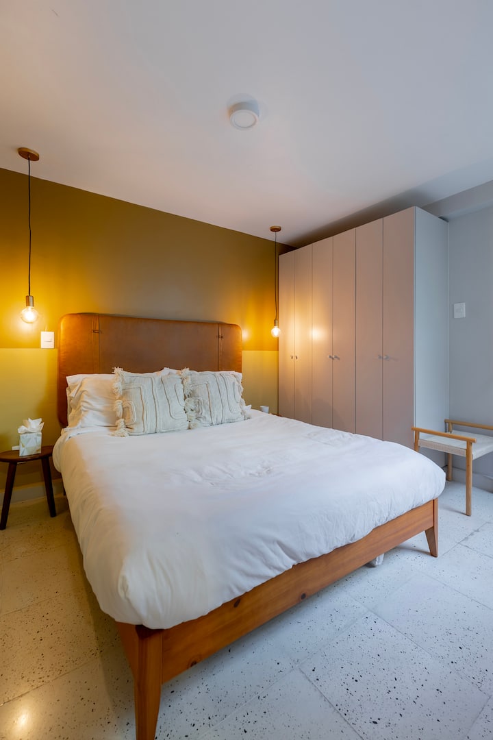 Recámara relajante con una lujosa cama de sábanas blancas frente a una pared de color oliva, enmarcada por lámparas colgantes y clósets empotrados. Los pisos de terrazo y el estilo suave crean un tranquilo refugio de hotel boutique en La Roma.