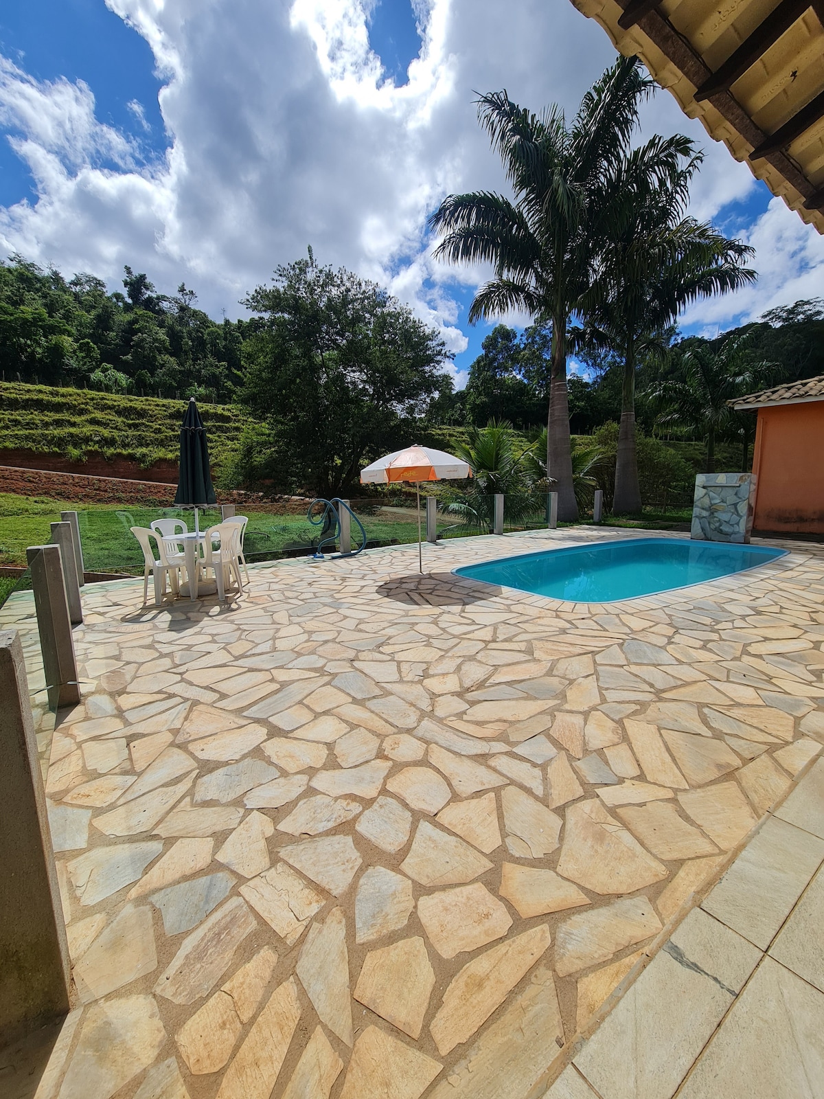 A sunlit patio features a rectangular pool surrounded by natural stone tiles. Two lounge chairs and a small table are positioned nearby, along with an umbrella providing shade. Lush greenery and palm trees frame the serene outdoor space, complemented by a clear blue sky.