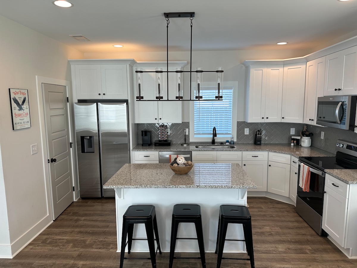 A modern kitchen showcases white cabinetry and a large central island with a granite countertop. Stainless steel appliances are neatly arranged, with a silver refrigerator and microwave visible. Three black stools are positioned at the island, providing seating for casual dining.