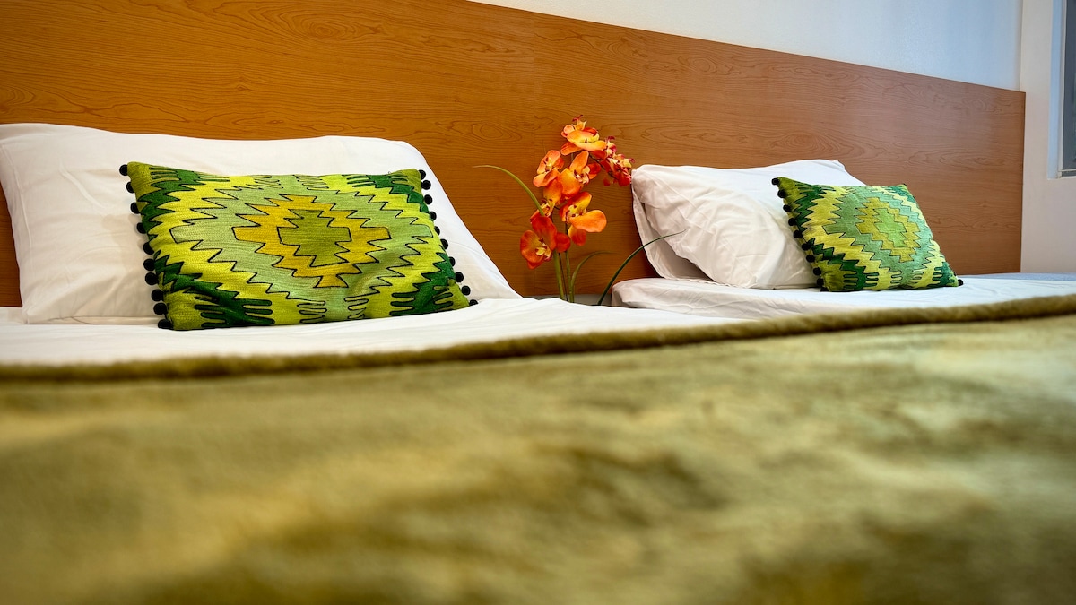 A minimalistic sleeping area is highlighted by two twin beds with white linens. Each bed features decorative green cushions. A small decorative plant with orange flowers adds a touch of color to the space, complementing the overall aesthetic.