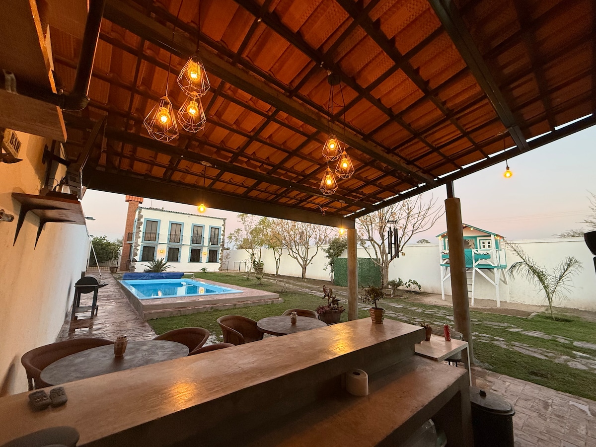 An outdoor terrace is displayed with overhead pendant lights providing illumination. A bar counter is visible, creating a social space. In the background, a swimming pool is surrounded by landscaping, with a partial view of the residential structure beyond.