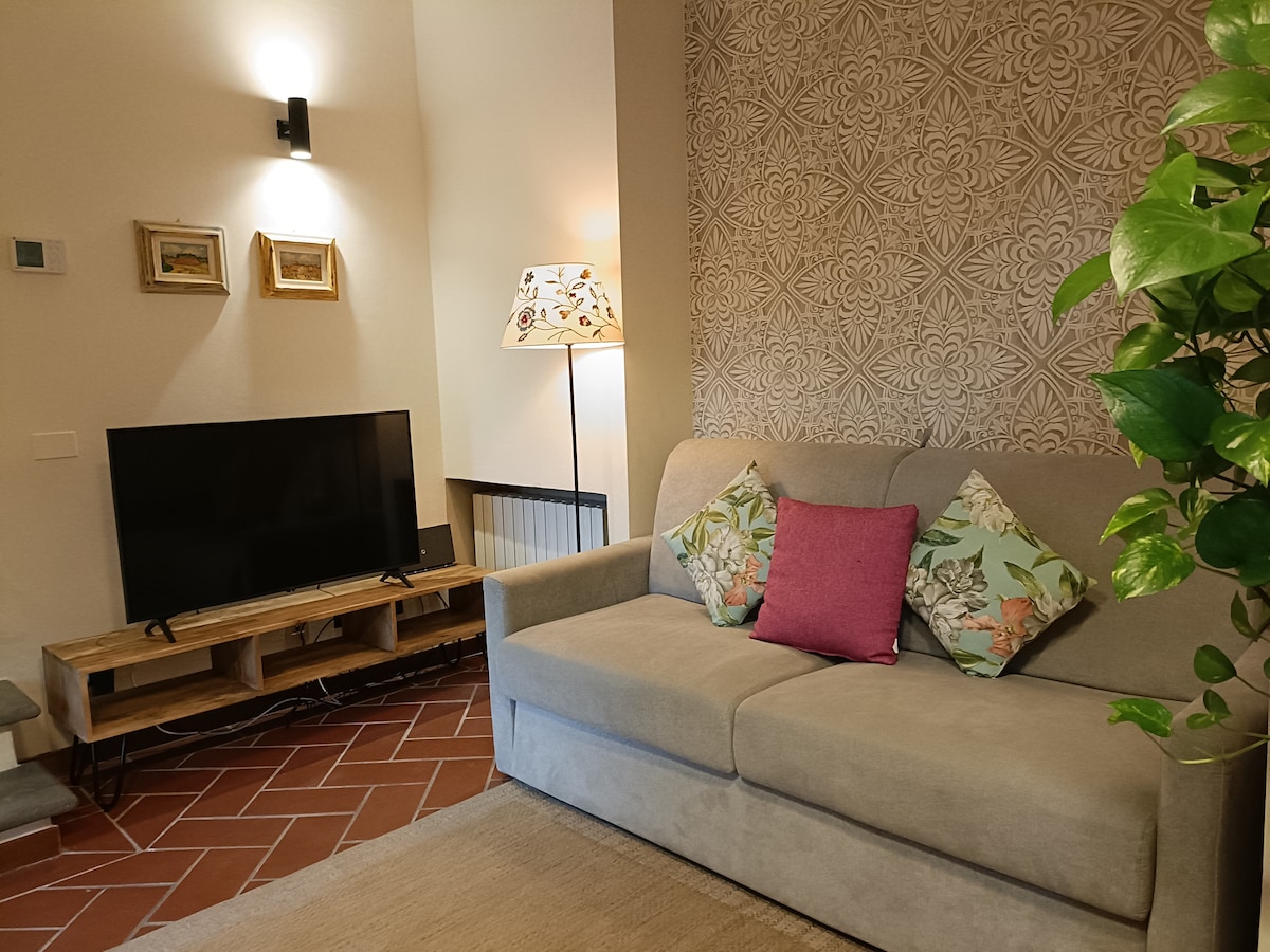 A comfortable living space features a plush sofa adorned with decorative pillows. A sleek wooden media console supports a television, while a decorative lamp adds a soft glow. Textured wallpaper and a small indoor plant contribute to the inviting atmosphere.