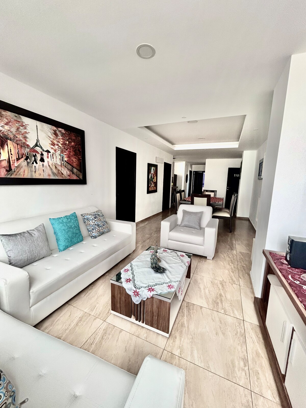 A bright living area features modern furnishings, including two white armchairs and a sleek coffee table at its center. Artwork on the walls adds a pop of color, contributing to a welcoming and well-lit atmosphere. Natural light fills the space, enhancing its openness.