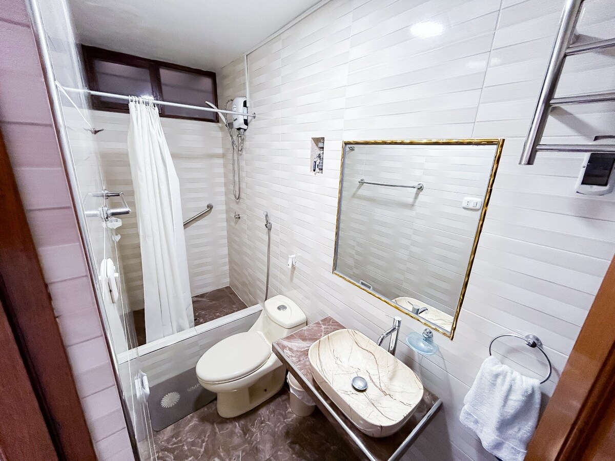 The bathroom features a shower area enclosed by a glass partition and has a modern sink set into a stone countertop. A toilet is located beside the sink, with grab bars for assistance. Light tiles on the walls enhance a clean and spacious feel.