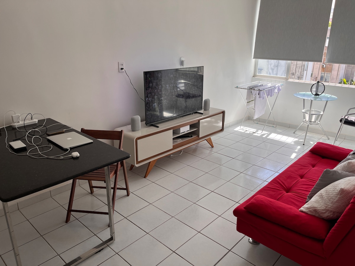 A bright living area is presented with a red sofa and a flat-screen TV mounted on a stylish media console. A small dining table with wooden chairs accompanies a folding iron board, while natural light floods in through the window, creating a cheerful and open feel.