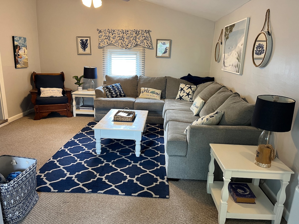A comfortable seating area is arranged with a spacious sectional sofa adorned with various cushions. A matching coffee table sits in the center, accompanied by decorative items. Light is provided by a ceiling fixture, and art pieces, including framed prints, are displayed on the walls.