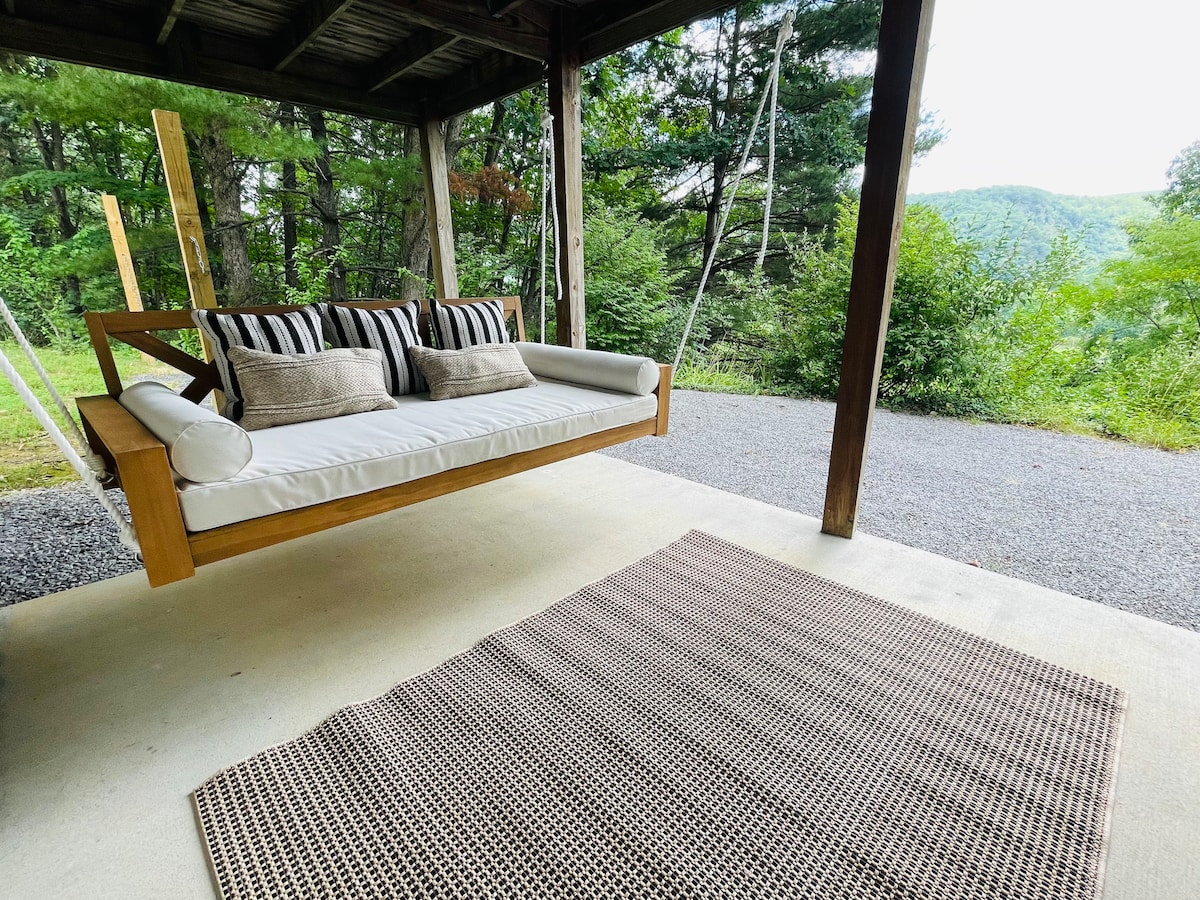 A comfortable swing bed is located on the covered patio, supported by wooden beams. Two striped cushions provide seating. A textured area rug lies underneath, and lush greenery and rolling hills are visible in the background, creating a serene outdoor atmosphere.