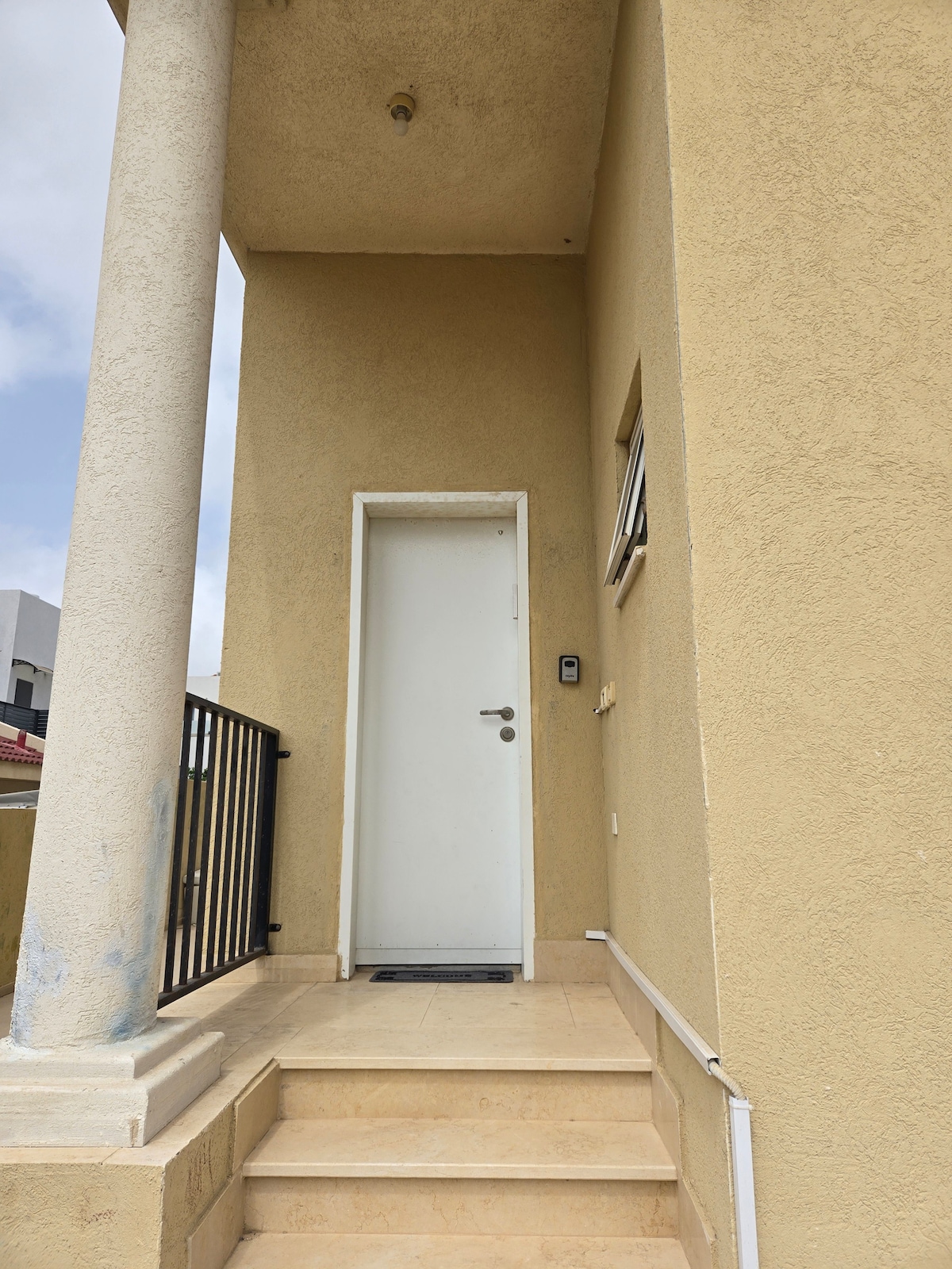 The entrance is marked by a simple white door flanked by light-colored walls. Two columns support the covered area above, while stone steps lead up to the doorway, providing access to the apartment's interior.
