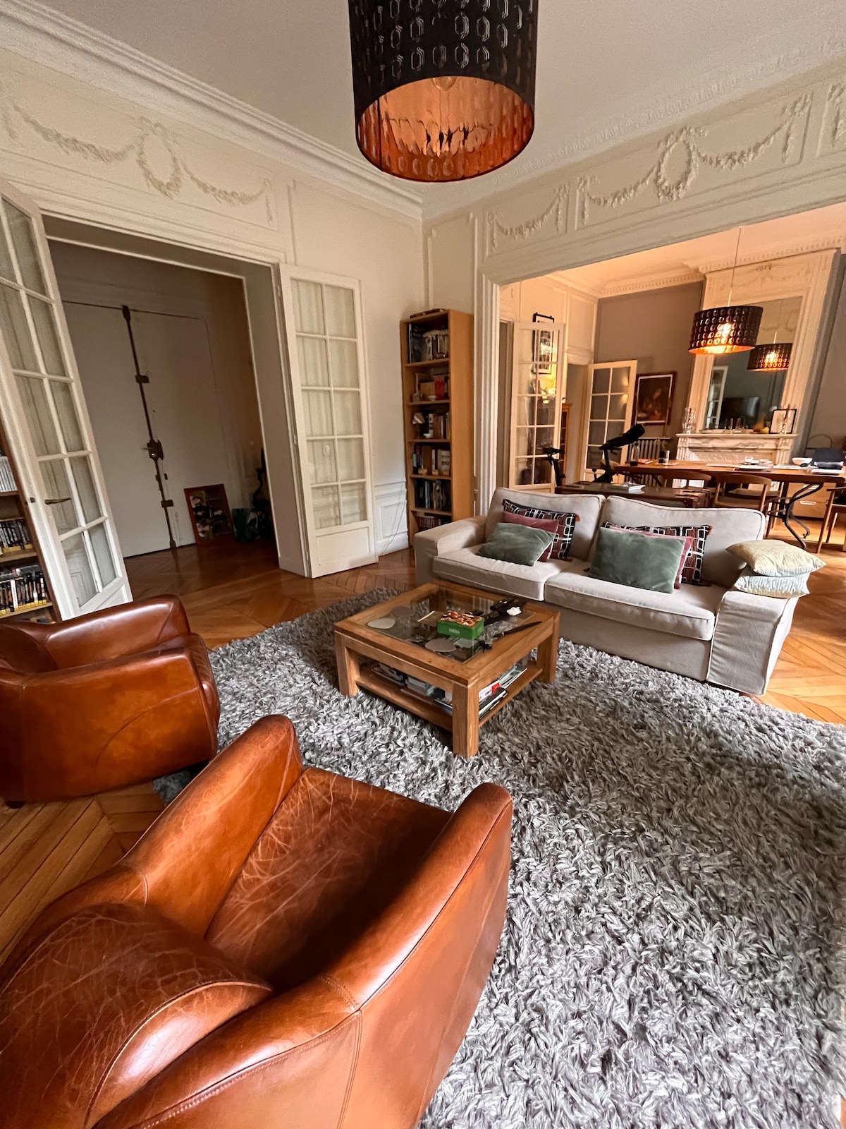 A cozy living area features a soft area rug beneath a light-colored sofa and two leather armchairs. A wooden coffee table is centrally placed, surrounded by bookshelves. The space opens to a dining area, separated by large double doors embellished with glass panels.