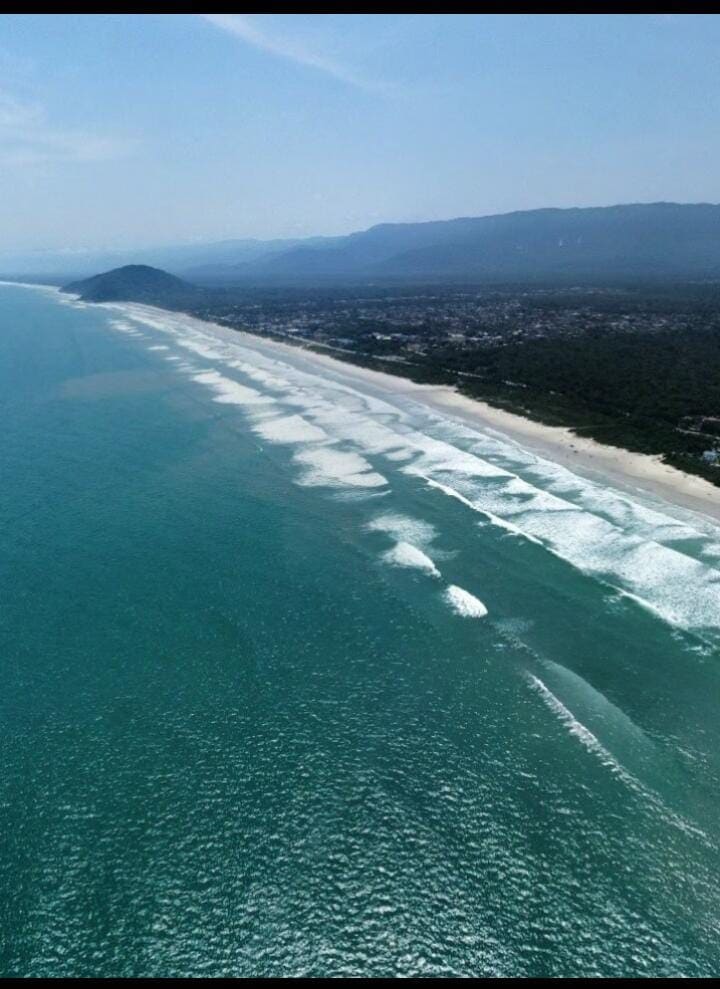 An aerial view captures a stretch of coastline featuring gently rolling waves meeting a sandy beach. The water reflects various shades of blue, while the surrounding landscape shows a mix of vibrant greenery and distant mountains under a clear sky.