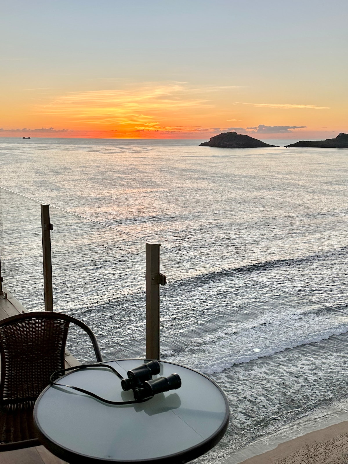 A private balcony offers an unobstructed view of the Pacific Ocean and a vibrant sunset. A round glass table, accompanied by a woven chair, is set for relaxation. Binoculars are placed on the table, inviting exploration of the scenic coastal landscape.