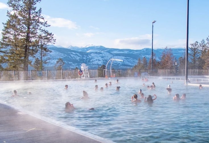 An outdoor hot spring pool is shown, surrounded by mist and mountains in the background. Numerous people can be seen enjoying the warm water, while trees and blue skies frame the scene. Spectacular mountain views are visible beyond the pool area.