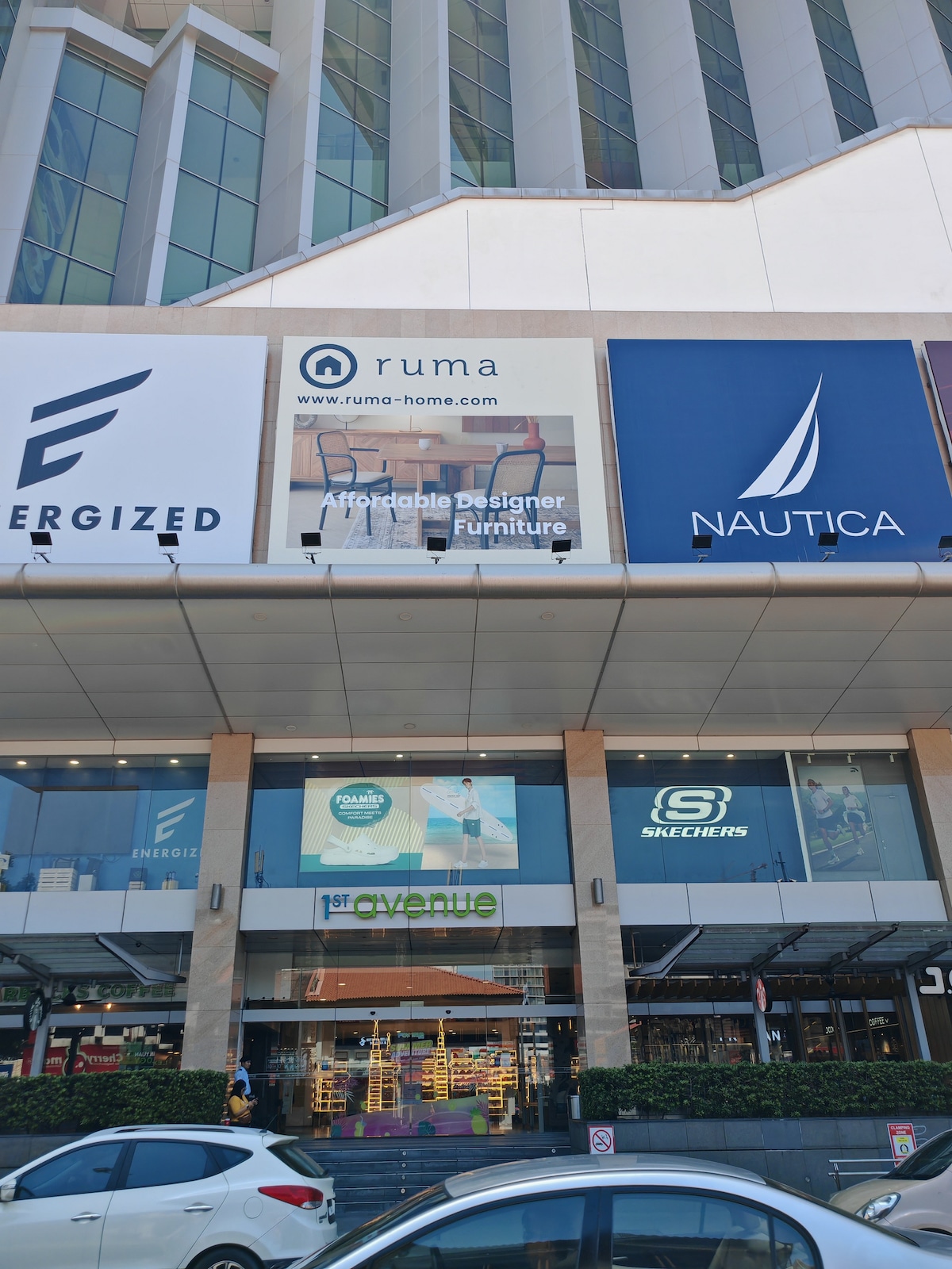The exterior of a modern building features a series of large advertisements for various brands. The ground level entrance is framed by sleek glass doors, with the signage for '1st Avenue' prominently displayed. A white car is parked in front of the building.
